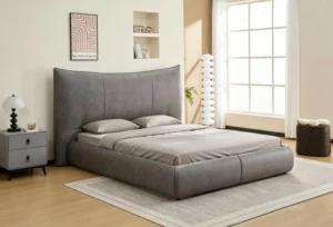 Grey upholstered bed with a tall curved headboard on a low upholstered base, in a pale room with a shuttered window, oak floor and a grey two-drawer nightstand beside it